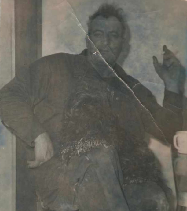 Black and white photo of man in overalls, sitting at a table, with a cigarette in hand and a dog on his lap